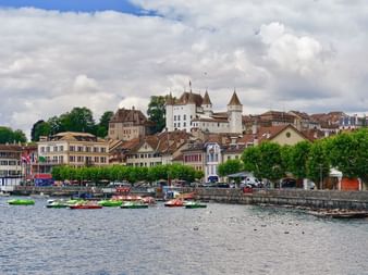 The town of Nyon on the lake with a view of the castle and pedalos in the water.