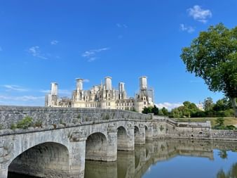 Vue du Château de Chambord dans la vallée de la Loire avec ses cheminées et tours distinctives, vu depuis un pont en pierre traversant une eau calme.