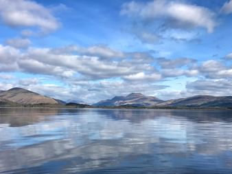 Loch Lomond in Schottland mit ruhigem Wasser, das sanfte Hügel und Berge unter einem bewölkten blauen Himmel perfekt spiegelt.