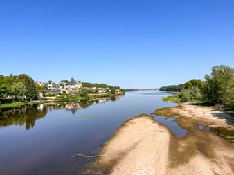 Vue large de la Loire avec bancs de sable et village de Candes-Saint-Martin sur la colline. Clocher visible parmi les bâtiments blancs sous ciel bleu.