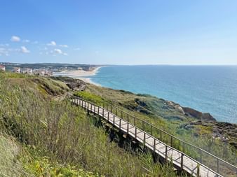 Ein Holzsteg auf einer begrünten Klippe mit Aussicht auf Häuser und Meer