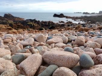 Strand auf Île de Bréhat bedeckt mit rosa und grauen Granitsteinen. Dunkle Felsen und ruhiges Meer im Hintergrund unter bewölktem Himmel.