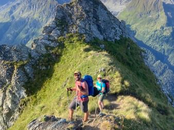 Reto und Nina wandern mit Rucksäcken auf einem schmalen Grasrücken am Col Termin, mit schneebedeckten Alpengipfeln im Hintergrund.