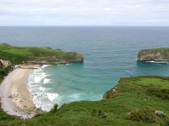 Abgeschiedener Sandstrand mit türkisfarbenem Wasser bei Llanes, umrahmt von grünen Klippen. Eine grasbewachsene Felsinsel steht im Meer.