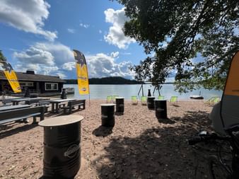 Eine Strandbar im Naherholungsgebiet Turku. Es hat einige Sitzgelegenheiten, Fässer und Liegestühle. Das Meer mit einer Schaukel befindet sich im Hintergrund. Rechts hat es einige Äste von einem Baum im Bild.