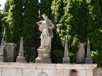Nepomuk-Statue auf Brücke in Salzburg Steinstatue des heiligen Johannes von Nepomuk auf einer barocken Brücke in Salzburg, umgeben von vier Obelisken und üppigen grünen Bäumen.