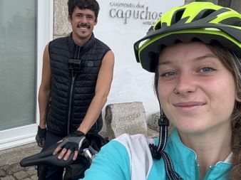 Zwei Radfahrer machen ein Selfie am Eingang des Hotels Convento dos Capuchos in Monção. Frau vorne trägt gelben Helm und türkisfarbene Jacke.