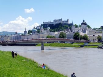 Blick auf Salzburg von der Salzach mit der historischen Altstadt, barocken Kirchen, der Festung Hohensalzburg auf dem Berg und einer Brücke.
