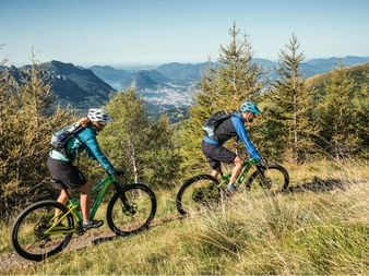 A mountain bike couple ride across a meadow. In the middle of the valley, surrounded by mountains, lies Lugano.