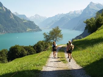 Deux randonneurs sur sentier de gravier à travers prairies vertes surplombant le lac des Quatre-Cantons turquoise entouré de montagnes alpines enneigées.