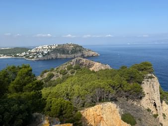 Felsige Küstenlandschaft in Nordkatalonien mit grüner Vegetation, blauem Mittelmeer und einer Hügelstadt auf einer Halbinsel unter klarem Himmel.