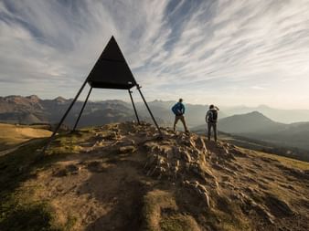 Two hikers stand on a mountain peak and gaze at the mountain landscape in front of them. Next to the hikers is a metal triangle with its tip pointing towards the sky.