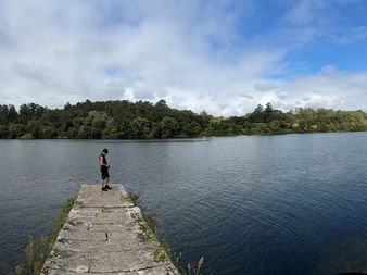 Person auf Steinpier am Rio Minho. Grüne Flussufer mit Bäumen auf beiden Seiten unter teilweise bewölktem Himmel.