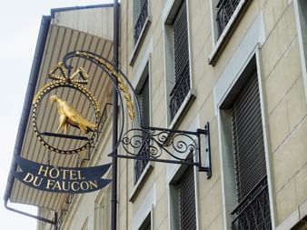 Verziertes Hotelschild des Hôtel du Faucon an einer historischen Gebäudefassade mit Fensterläden in Freiburg. Das Schild zeigt einen goldenen Falken.