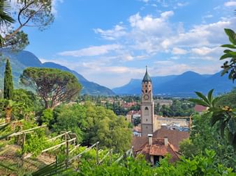 Vue panoramique de Merano depuis le Tappeinerweg montrant le clocher historique, les bâtiments aux toits rouges et les montagnes alpines.