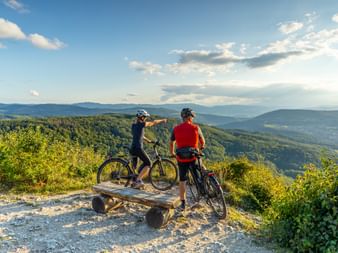Zwei E-Biker auf einer Holzbank am Aussichtspunkt Gempen in der Region Basel mit Blick auf bewaldete Hügel und Täler unter blauem Himmel.