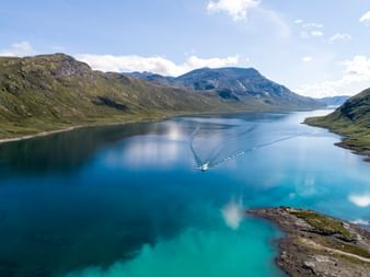 Luftaufnahme eines Bootes auf dem türkisfarbenen Bygdin-See in Norwegen, umgeben von grünen Bergen unter blauem Himmel mit weißen Wolken.