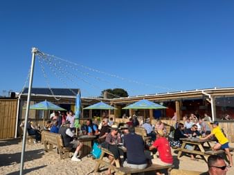 Gut besuchtes Strandcafé in Cornwall mit Holzpicknicktischen im Sand. Gäste sitzen unter bunten Sonnenschirmen bei blauem Himmel.