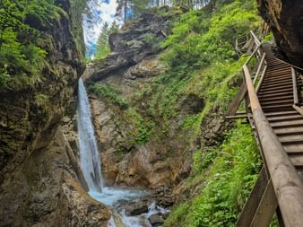 Wasserfall in der Raggaschlucht mit Holzsteg auf der rechten Seite. Üppige grüne Vegetation bedeckt die moosbewachsenen Felsen.