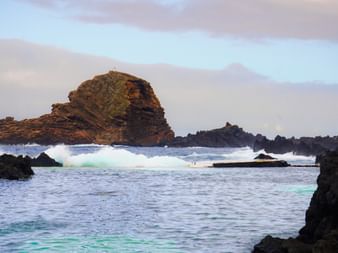 Wellen brechen sich an dunklen Vulkanfelsen auf Madeira, Portugal. Eine große braune Felsformation erhebt sich aus dem Ozean mit Bergen im Hintergrund.