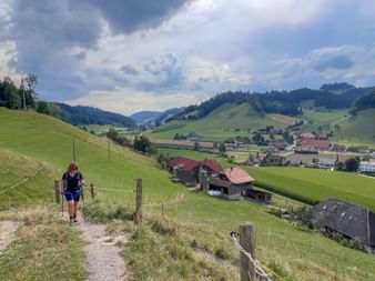Eine Wanderin mit Trekkingstöcken steigt einen steilen Pfad hinauf, umgeben von grünen Hügeln und einem malerischen Tal im Hintergrund.