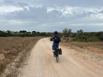 Radfahrerin mit Helm und Rucksack auf Feldweg durch trockene Landschaft in der Algarve, mit bewölktem Himmel und entferntem Dorf.
