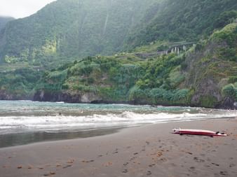 Dunkler Sandstrand auf Madeira mit weißen Wellen, rotem Surfbrett am Ufer und steilen grünen Bergen mit terrassierten Hängen und Brücke im Hintergrund.