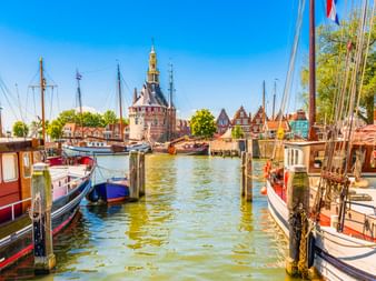 Bateaux traditionnels amarrés dans le port de Hoorn avec tour historique et maisons à pignons sous ciel bleu. Poteaux en bois bordent le canal.