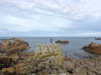 Person auf felsigem Küstenvorsprung bei Plougrescant, Bretagne. Geschichtete Felsformationen erstrecken sich ins ruhige Meer unter bewölktem Himmel.