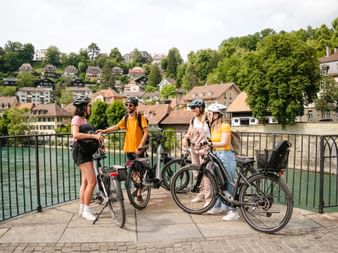 Vier Radfahrer mit Helmen und Rädern stehen auf Steinbrücke mit Blick auf türkisfarbene Aare in Berns historischer Altstadt mit traditionellen Gebäuden.