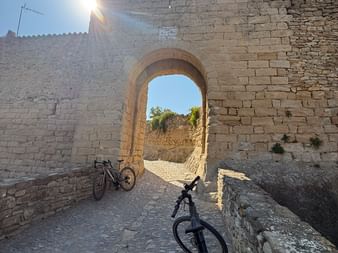 Zwei Fahrräder stehen an einem gewölbten Steintor in einer mittelalterlichen Festungsmauer. Sonnenlicht strömt durch und erzeugt Linsenreflexe.