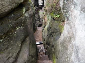 Steile Treppe durch eine enge Felsspalte zwischen moosbewachsenen Felswänden auf dem Malerweg. Ein Wanderer ist oben sichtbar.