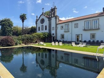 Historisches weißes Convento dos Capuchos mit Kapelle und Glockenturm neben rechteckigem Pool. Liegestühle auf grünem Rasen unter blauem Himmel.