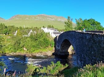Steinbogenbrücke über einen Fluss bei Bridge of Orchy, Schottland, mit weißen Gebäuden und grünen Hügeln unter blauem Himmel.