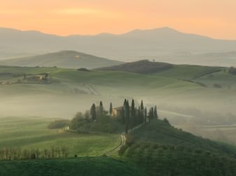 Collines verdoyantes de Toscane au lever du soleil avec brume matinale, villa sur colline entourée de grands cyprès et chemin de terre.