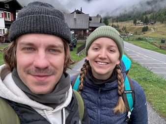 Zwei Wanderer mit Rucksäcken im Dorf Gruben am Alpenpässeweg. Schneebedeckte Berge und Nebel hinter traditionellen Alpenhäusern sichtbar.