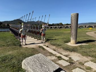 Reihe römischer Soldatenstatuen mit Speeren neben einer Steinsäule, im Hintergrund historische Bogenbrücke über den Rio Lima.