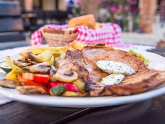Gegrilltes Schweinekotelett mit Kräuterbutter auf weißem Teller, serviert mit gegrilltem Gemüse, Pilzen und Pommes. Brotkorb im Hintergrund.
