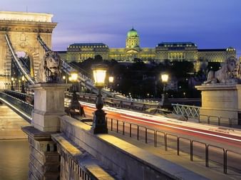 Beleuchtete Budapester Kettenbrücke bei Dämmerung mit steinernen Löwenstatuen über der Donau. Königspalast mit grüner Kuppel auf dem Hügel.