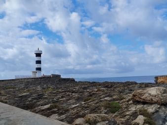 An der Küste befindet sich ein schwarz-weiss-gestreifter Leuchtturm. Dahinter ist das Mittelmeer in Mallorca.