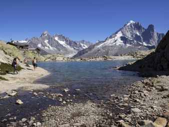 Kristallklarer Bergsee mit Wanderern am felsigen Ufer, umgeben von schneebedeckten Mont Blanc Gipfeln unter blauem Himmel.