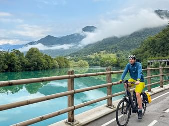 Radfahrer mit gelben Taschen auf einer Brücke mit Holzgeländer über türkisfarbenem Fluss, bewaldete Berge und tiefe Wolken im Hintergrund.