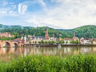 Panoramablick auf Heidelberg mit der roten Sandstein Alten Brücke über den Neckar, bunten historischen Gebäuden am Flussufer und dem Heidelberger Schloss am bewaldeten Hang.