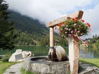 Ein mit Geranien bepflanzter, runder Brunnen vor dem See.