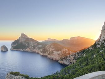 Dramatic sunset view over Mallorca's rugged coastline with limestone cliffs, calm blue sea, and mountains bathed in golden light.