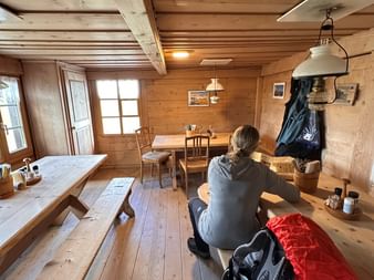 Wanderin sitzt auf Bank in Holz-Alphütte mit Rucksack und roter Isomatte. Rustikales Interieur mit Holztischen, Bänken und Fenstern.