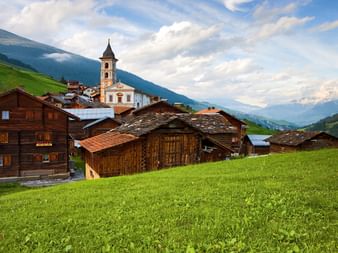 Alpendorf Vrin im Val Lumnezia mit traditionellen Holzchalets, weißer Kirche mit Glockenturm, umgeben von grünen Wiesen und Bergen.