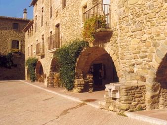 Mittelalterliche Steingebäude mit gewölbten Türen und Balkonen in einem nordkatalanischen Dorf. Ein Steinbrunnen steht im sonnigen Innenhof.
