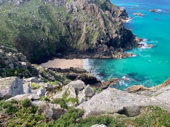 Luftaufnahme der dramatischen Felsküste Cornwalls mit steilen Klippen, einem kleinen Sandstrand und türkisfarbenem Atlantikwasser mit sichtbaren Felsen.