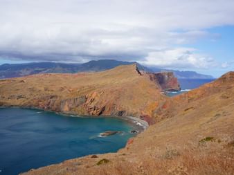 Felsige Küstenklippen in rotbraunen Tönen umgeben eine türkisfarbene Bucht auf Madeira, mit Bergen über dem Meer unter bewölktem Himmel.